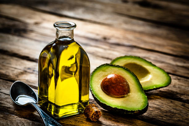 Vegan food: extra virgin avocado oil in a glass bottle shot on rustic wooden table. Sliced organic avocado and a spoon filled with oil are beside the bottle. Predominant colors are yellow and green. 